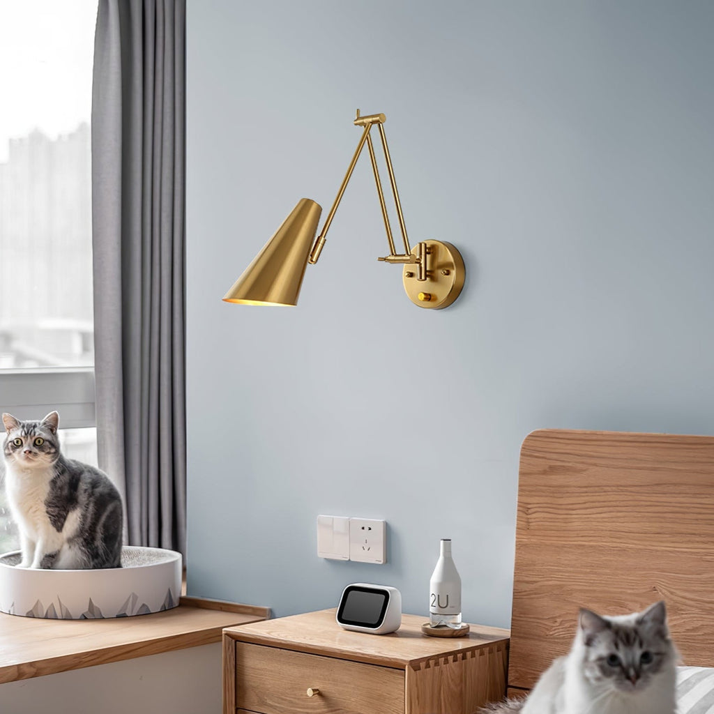 A Clérachic gold wall light with a conical shade on a pale blue wall above a wooden bedside table, with two cats nearby.