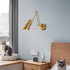 A Clérachic gold wall light with a conical shade on a pale blue wall above a wooden bedside table, with two cats nearby.