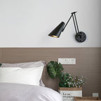 Black conical-shade Clérachic wall light on a white wall above a bed with a taupe headboard, with a large leafy plant at the side.