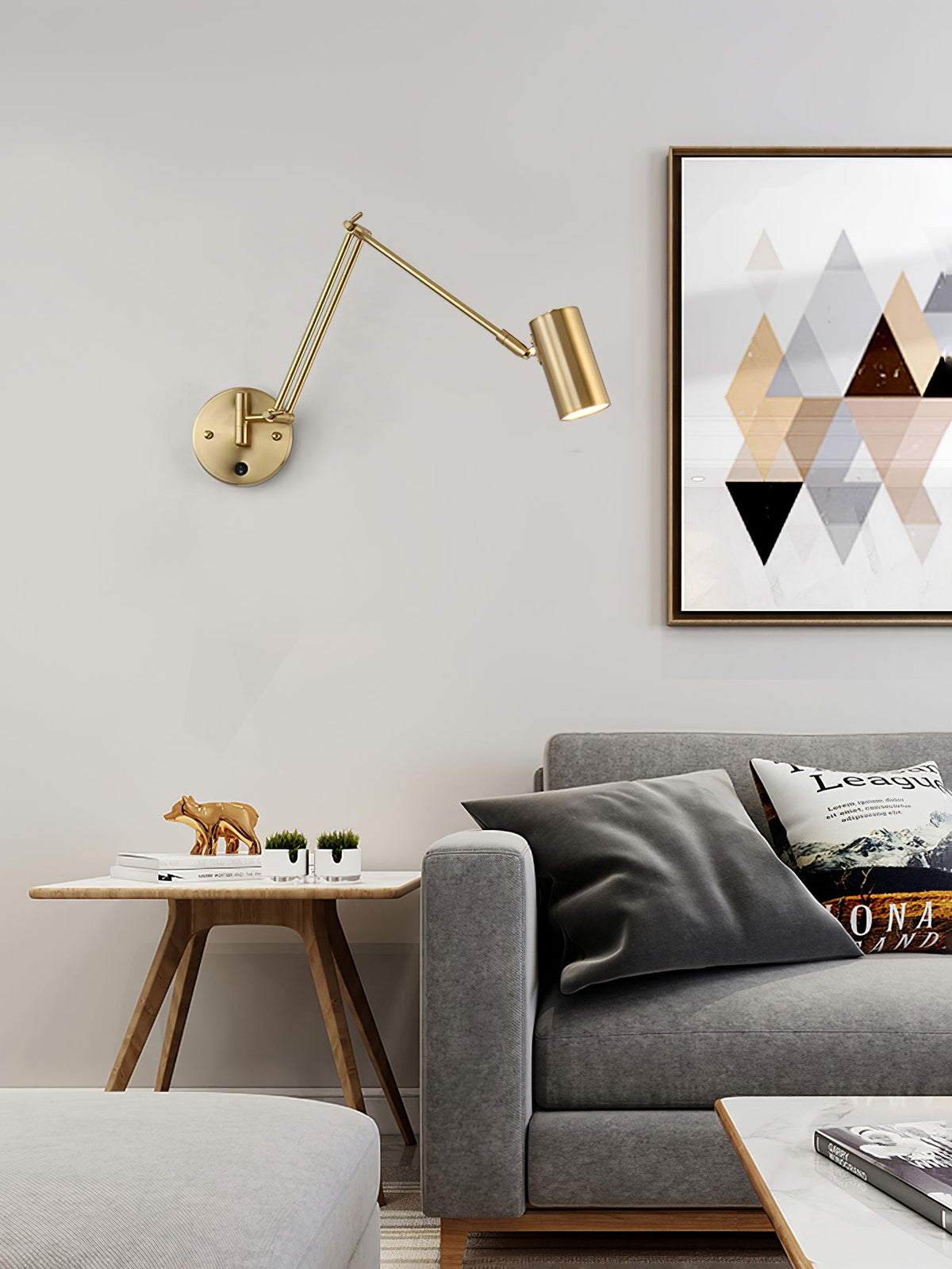 Gold cylinder-head Clérachic swing-arm wall light on a grey wall beside a framed geometric print, above a grey sofa and a wooden side table.