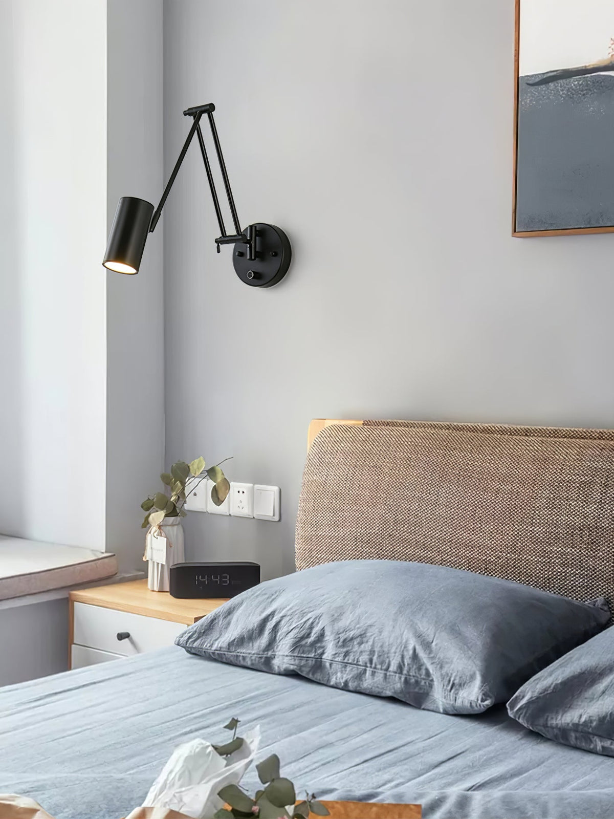 A black Clérachic swing-arm wall light with a cylinder head beside a bed with blue-grey bedding and a woven headboard.