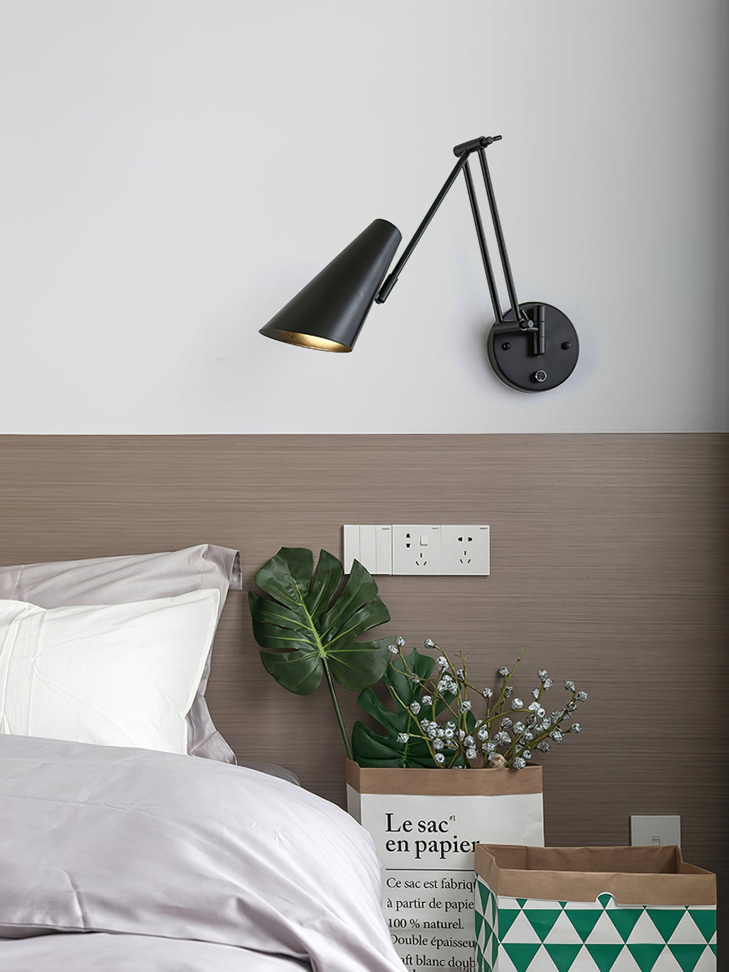 Clérachic black wall light with a conical shade and a folding arm, above a bed with a taupe headboard and a potted plant.