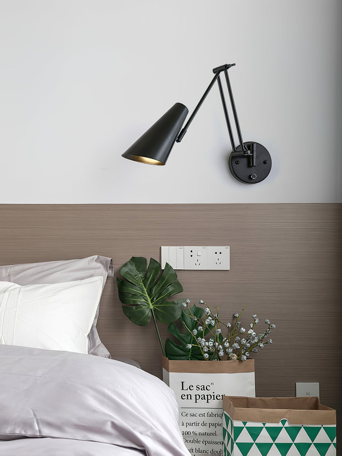 Clérachic black wall light with a conical shade and a folding arm, above a bed with a taupe headboard and a potted plant.