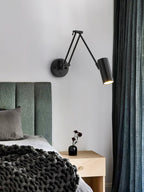A black Clérachic swing-arm wall light with a cylinder head, reaching over a bedside table with a vase, beside a green headboard and dark curtains.