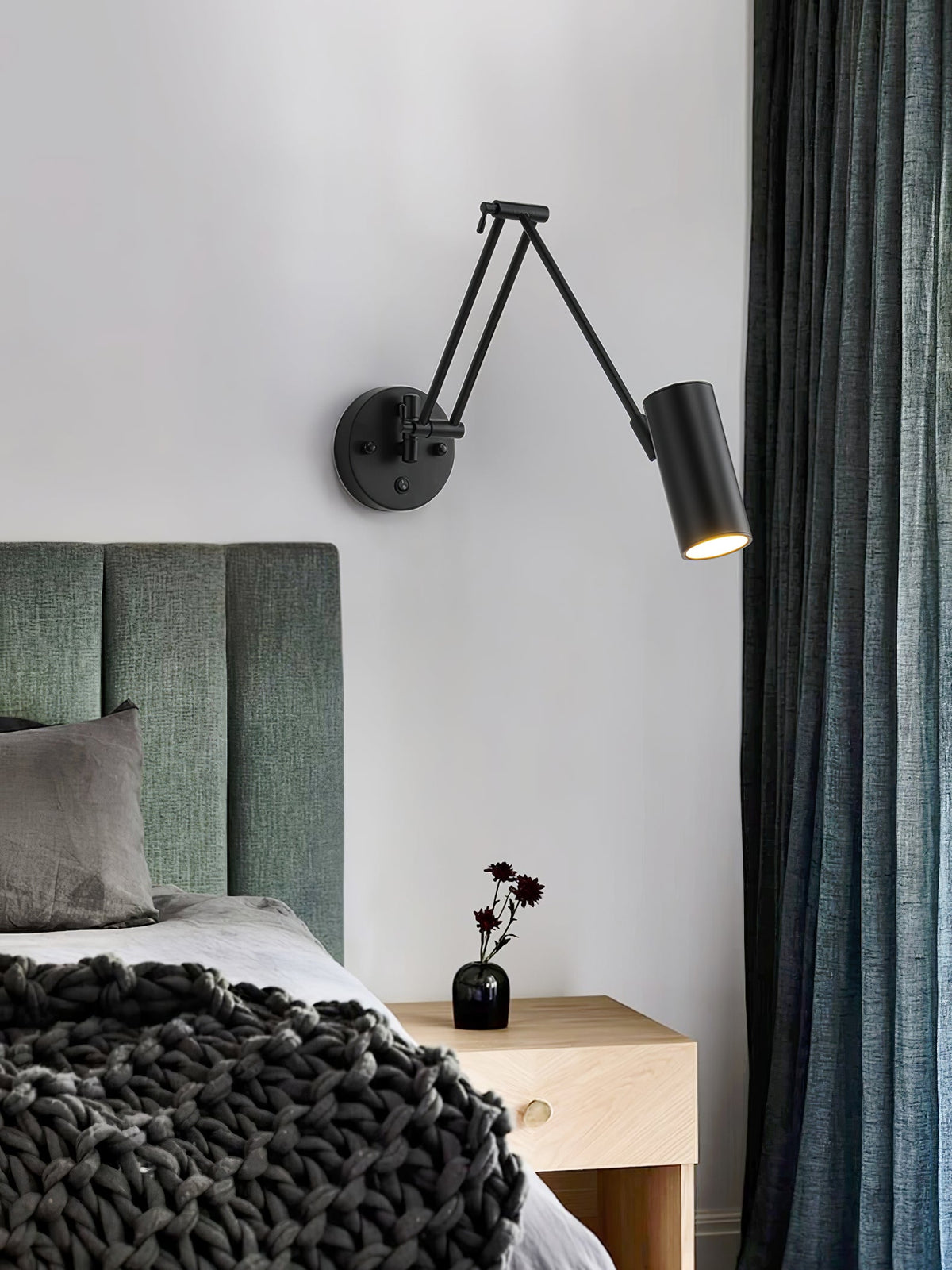 A black Clérachic swing-arm wall light with a cylinder head, reaching over a bedside table with a vase, beside a green headboard and dark curtains.