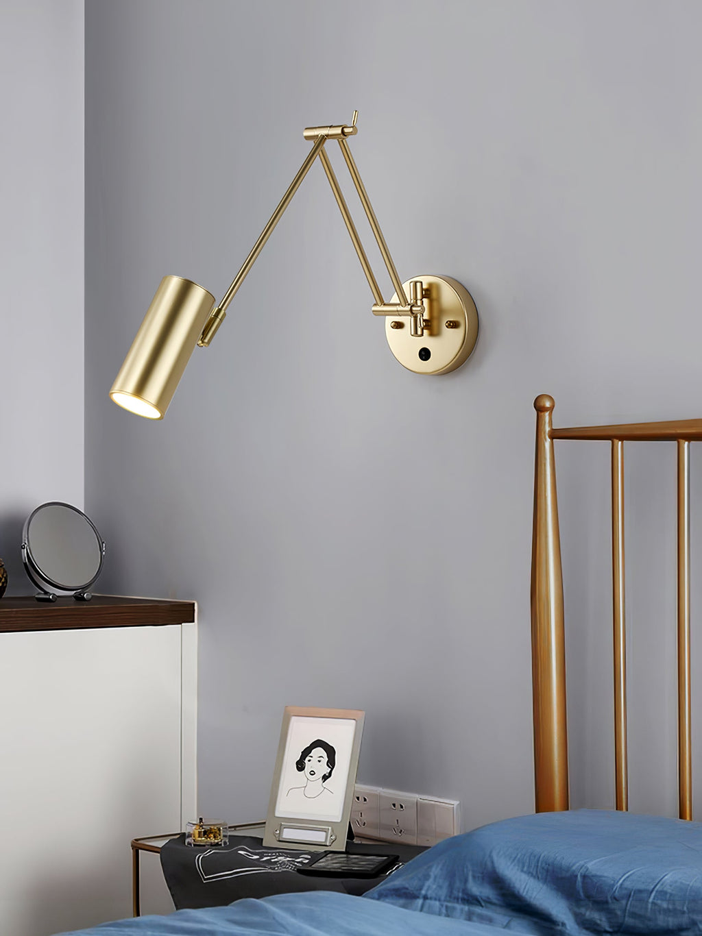Clérachic gold wall light with a cylinder head on a grey-blue wall above a bedside table with a framed portrait, beside a brass bed frame.