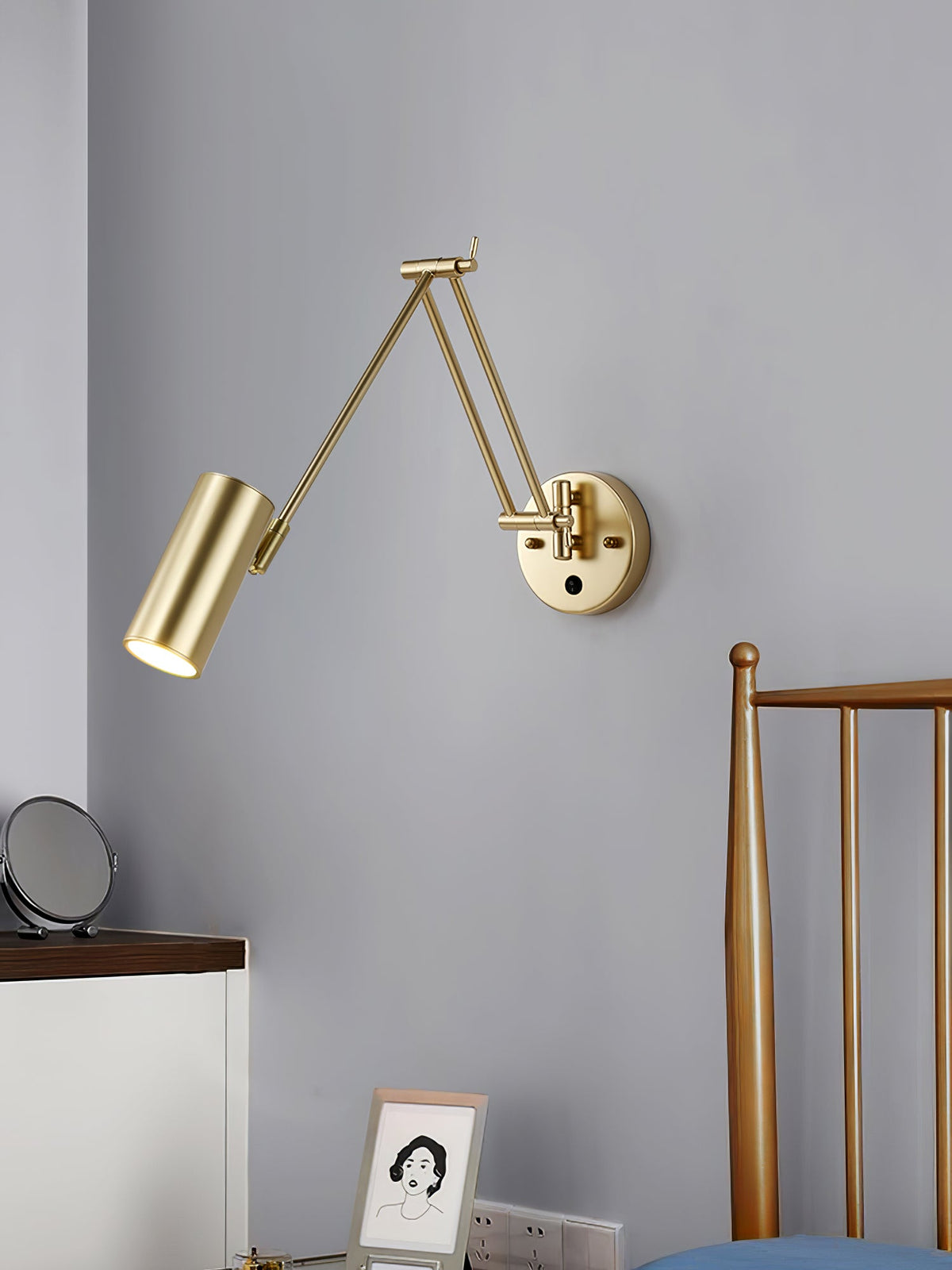 The gold cylinder-head Clérachic wall light on a grey-blue wall, with the arm folded, above a bedside table and a brass bed frame.