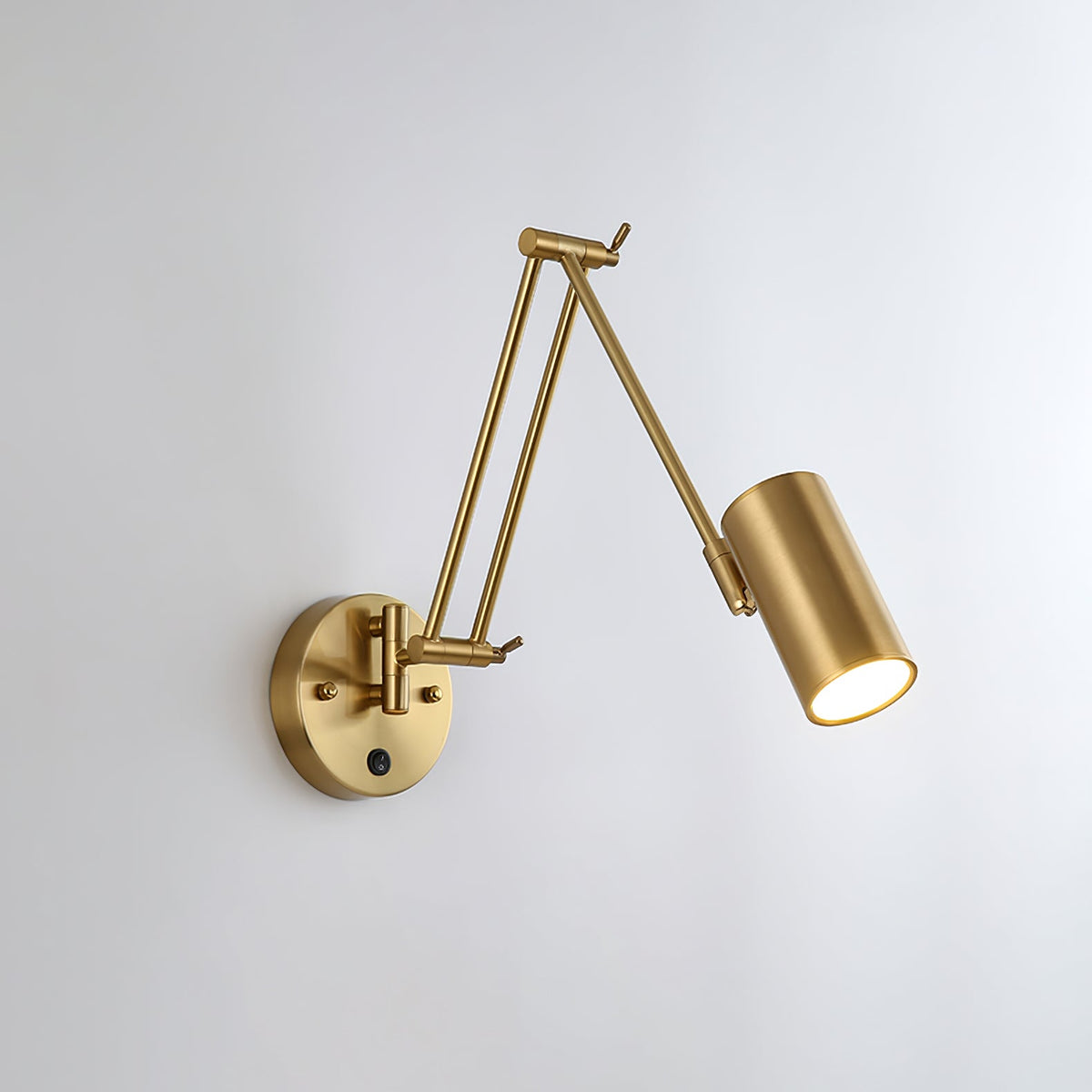 Clérachic gold wall light with a cylinder head angled down to the right on a folded arm, on a pale wall.