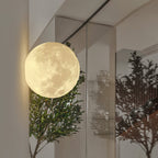 Round Clérachic moon wall light glowing beside a glass partition, with potted olive trees reflected in the panels.