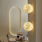 Two round Clérachic moon wall lights, one above the other, beside a lit oval niche and a small café table with a pastry.