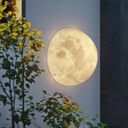 Round Clérachic moon wall light lit against an exterior wall at dusk, with yellow-leaved branches in front.