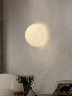 Round Clérachic moon wall light on a mottled plaster wall above a cream armchair and a dried branch in a vase.