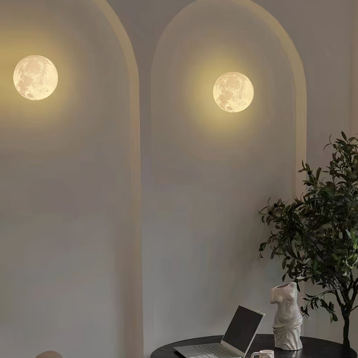 Two round Clérachic moon wall lights set in arched plaster recesses, with a plant and a laptop on a table below.