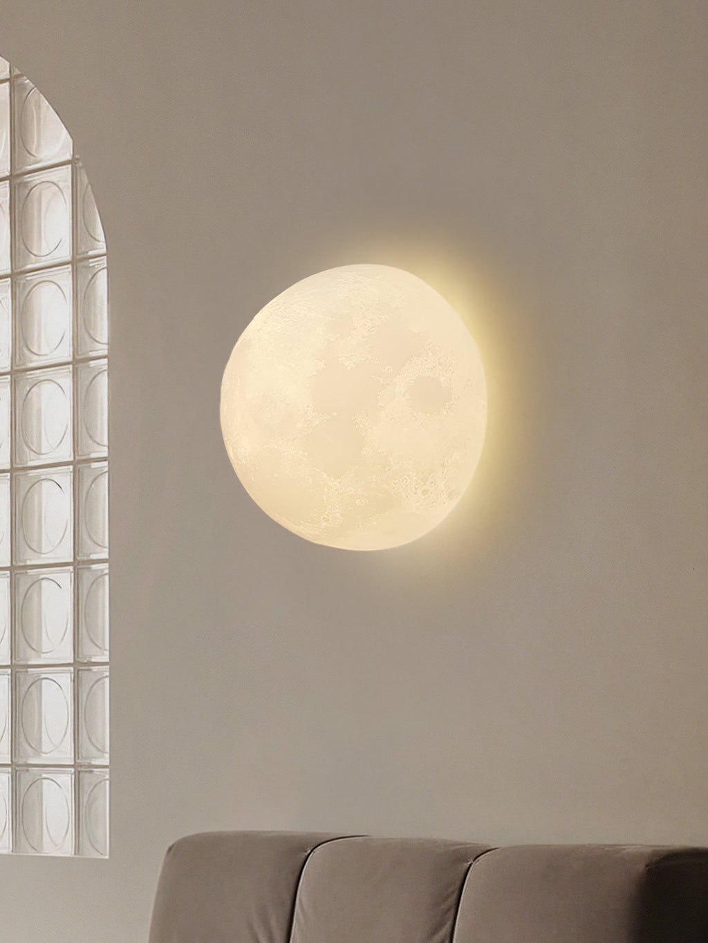 Round Clérachic moon wall light on a beige wall beside an arched glass-block window and the back of a grey sofa.