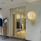 Round Clérachic moon wall light on a white shopfront wall beside a glass door and clothing mannequins.