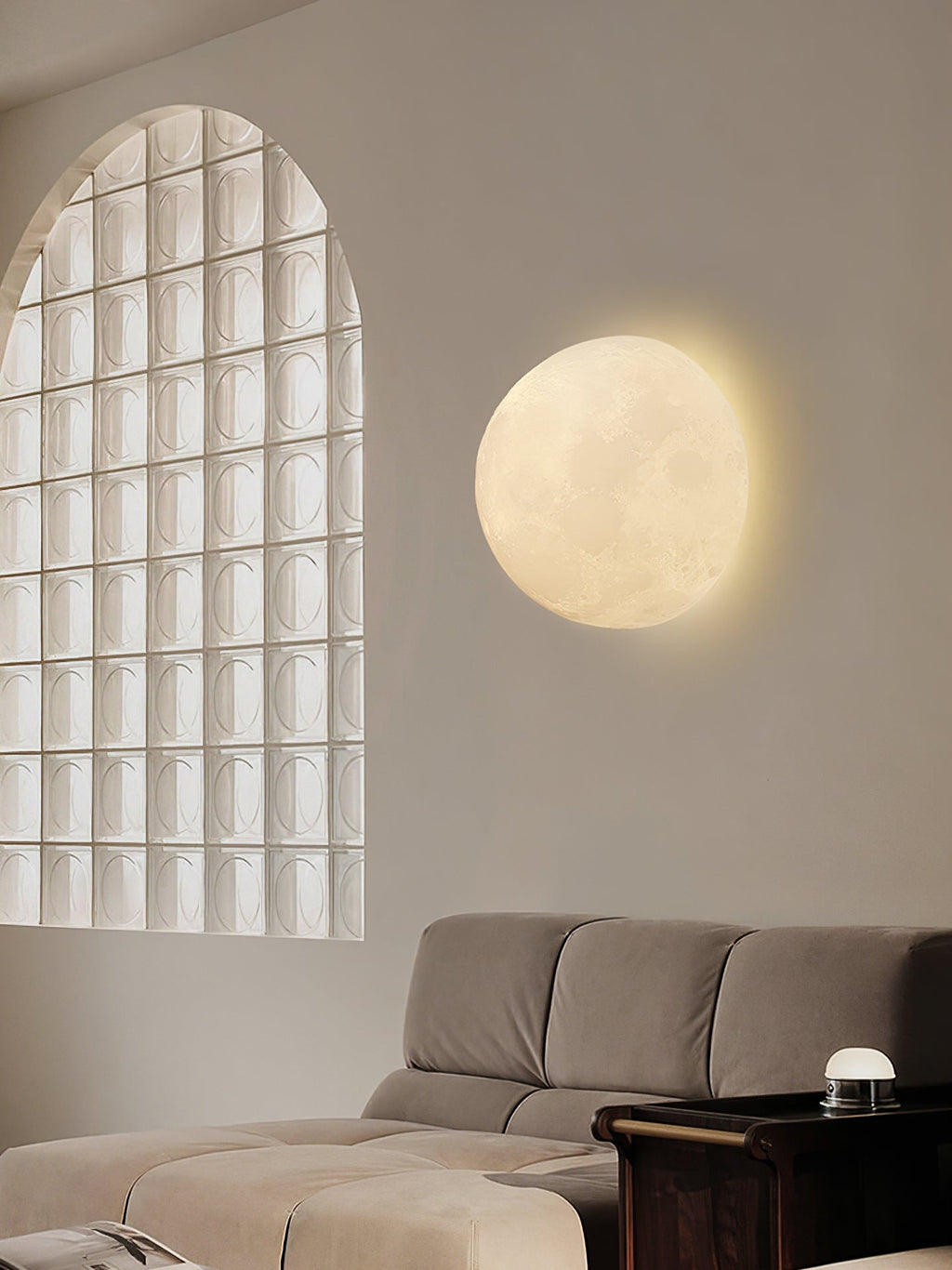 Round Clérachic moon wall light beside an arched glass-block window, above a grey sofa and a small dark side table.