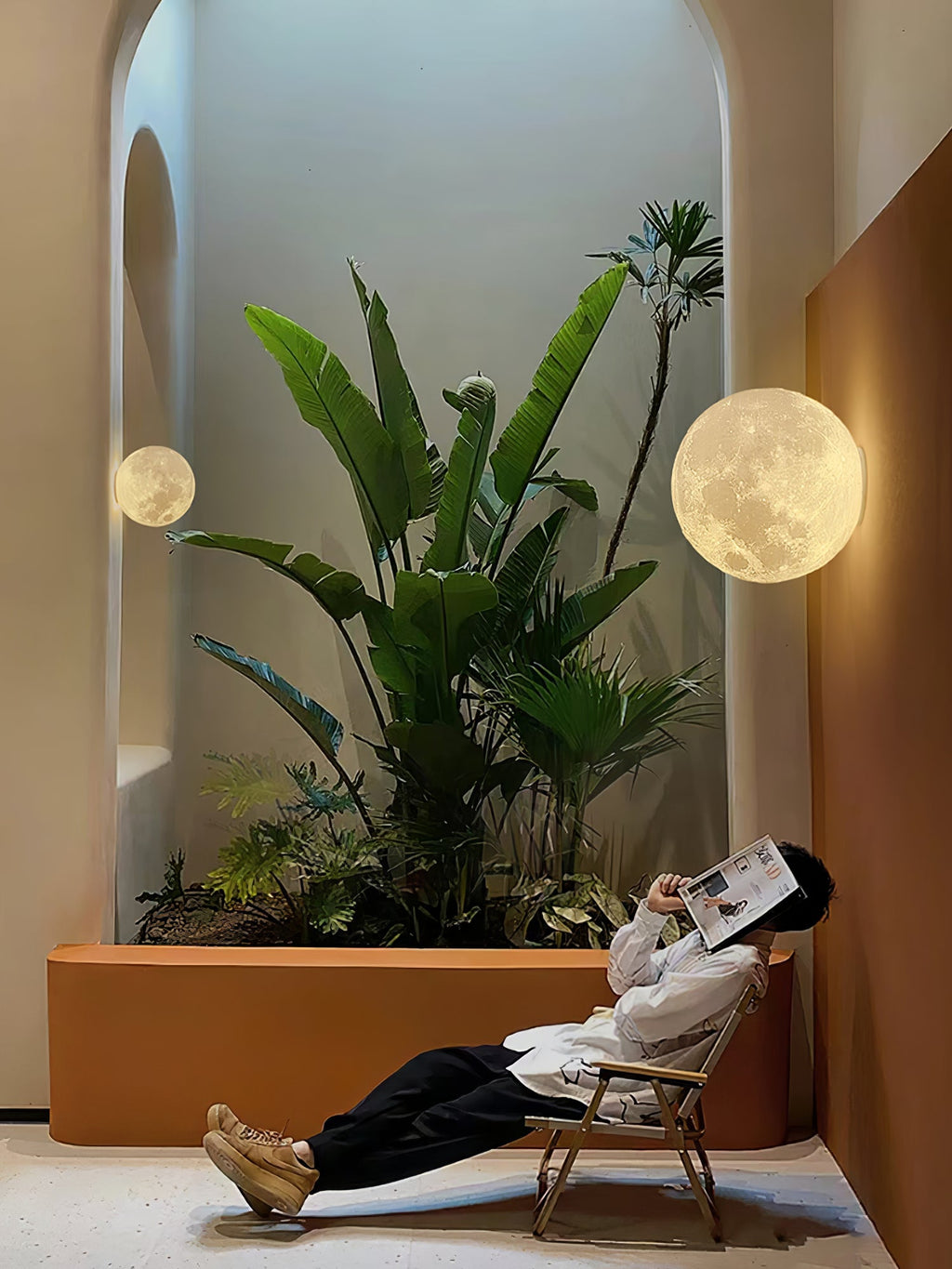 Round Clérachic moon wall light on a terracotta wall above a planter of large-leaved plants, with a person reclining below.