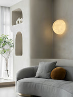 Round Clérachic moon wall light above a grey curved sofa, beside arched wall niches and a tall plant in a glass vase.