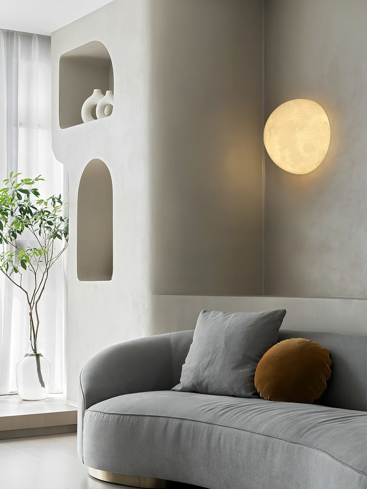 Round Clérachic moon wall light above a grey curved sofa, beside arched wall niches and a tall plant in a glass vase.