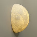 Angled view of a Clérachic half-moon wall light, showing the pitted lunar surface lit from within against a taupe wall.