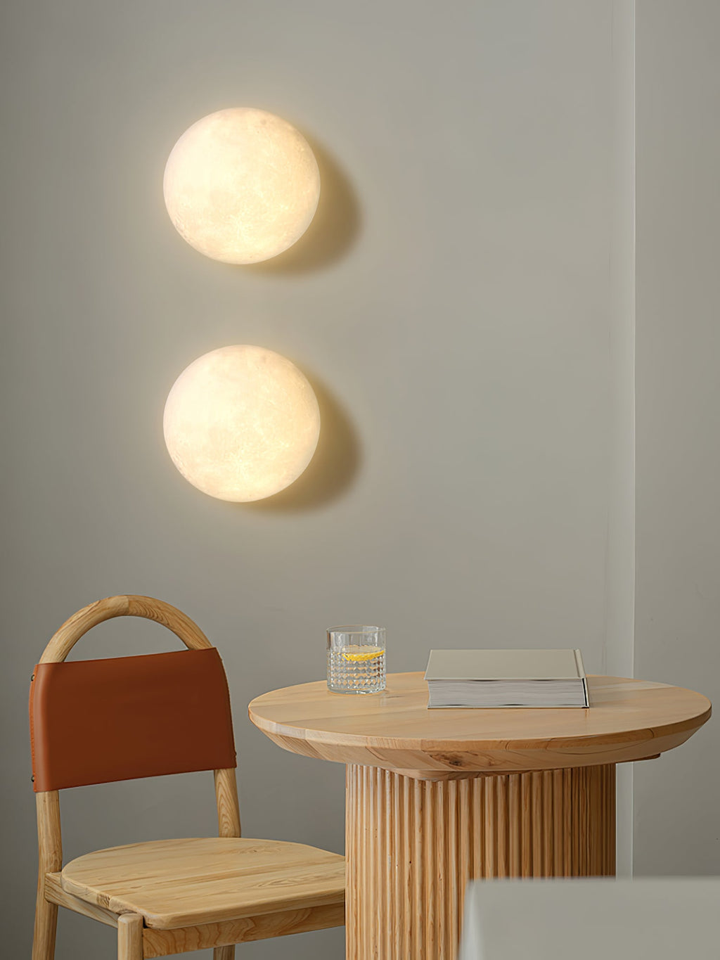 Two round Clérachic moon wall lights above a round wooden table with a ribbed base, a rattan chair and a glass of water.