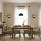 A pair of small Clérachic wall lights flank a window in a dining room with panelled walls, a wooden table and green spindle chairs.