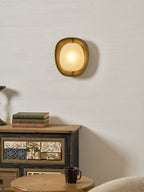 On a textured cream wall, a lit Clérachic wall light glows above a painted sideboard with a stack of books and a coffee mug.