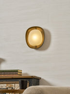 This lit Clérachic wall light has a rounded bronze rim and a warm frosted shade, with a sideboard and soft armchair below.