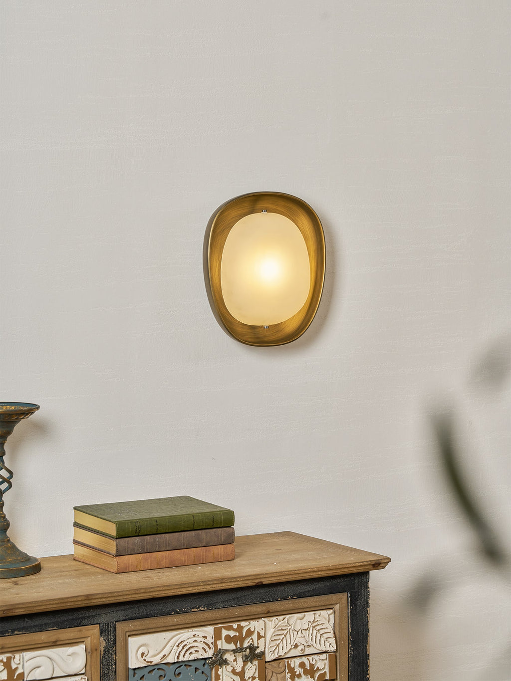 A lit Clérachic wall light on a plaster-textured wall, with a stack of books on a wooden sideboard below it.