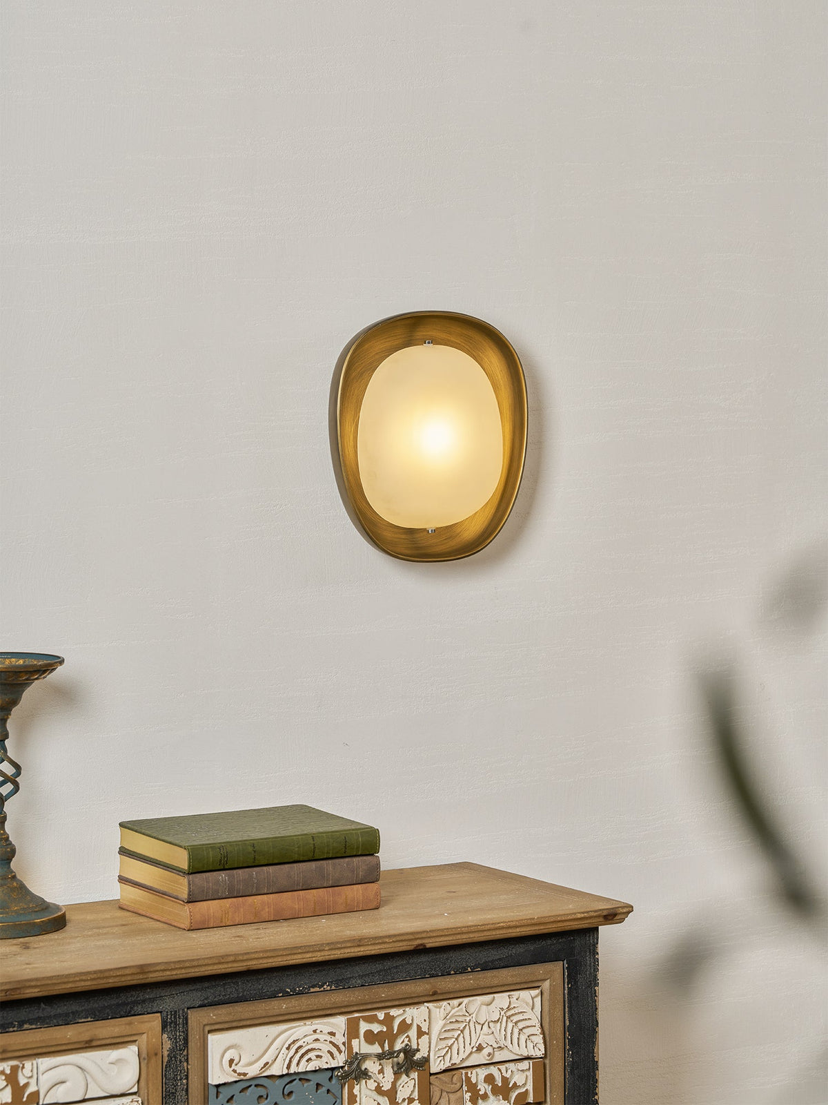 A lit Clérachic wall light on a plaster-textured wall, with a stack of books on a wooden sideboard below it.