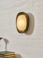 Seen at a slight angle, a lit Clérachic wall light shows its deep bronze bowl and glowing frosted oval shade on a textured wall.