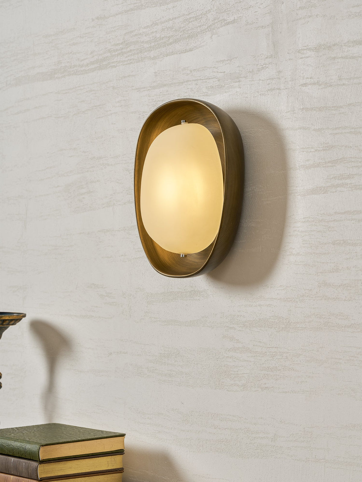 Seen at a slight angle, a lit Clérachic wall light shows its deep bronze bowl and glowing frosted oval shade on a textured wall.