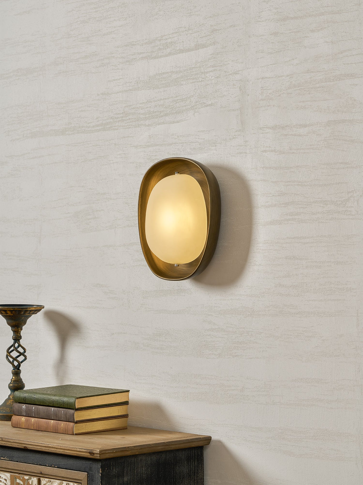 This Clérachic wall light glows warm on a plaster wall, above a sideboard with books and an ornate metal candle stand.