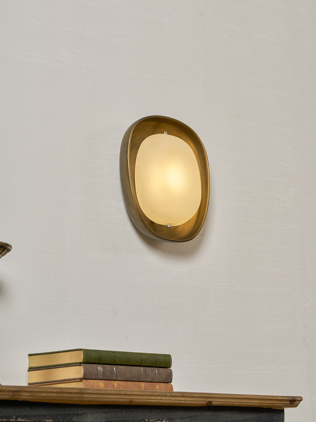 A warm-lit Clérachic wall light, angled slightly, on a pale wall, with a stack of books on a wooden shelf below.