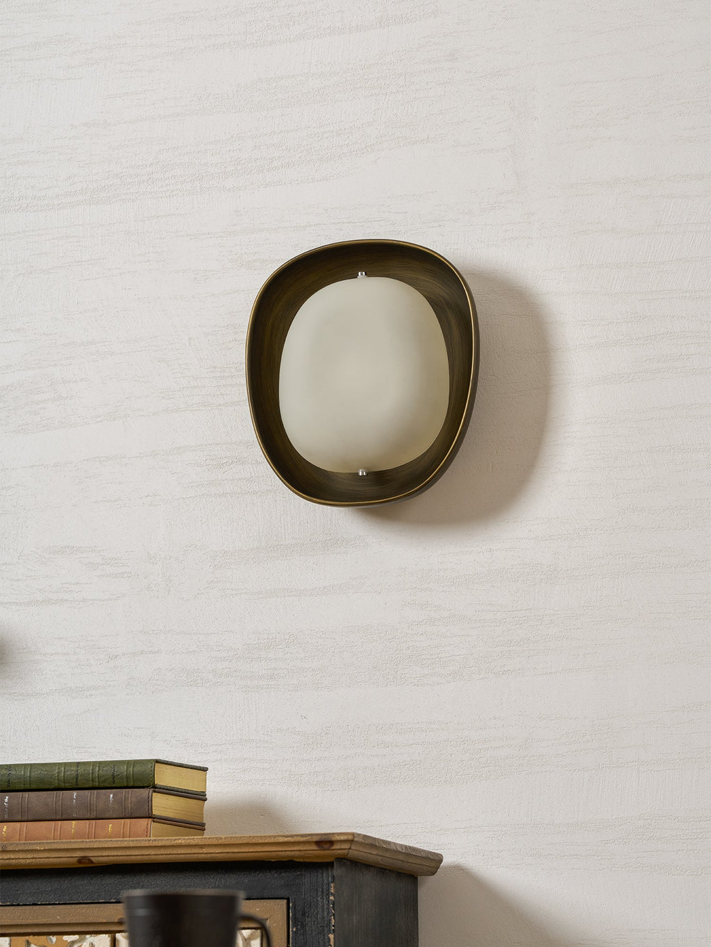 This Clérachic wall light, switched off, shows a frosted white shade inside a rounded bronze rim, on a textured pale wall.