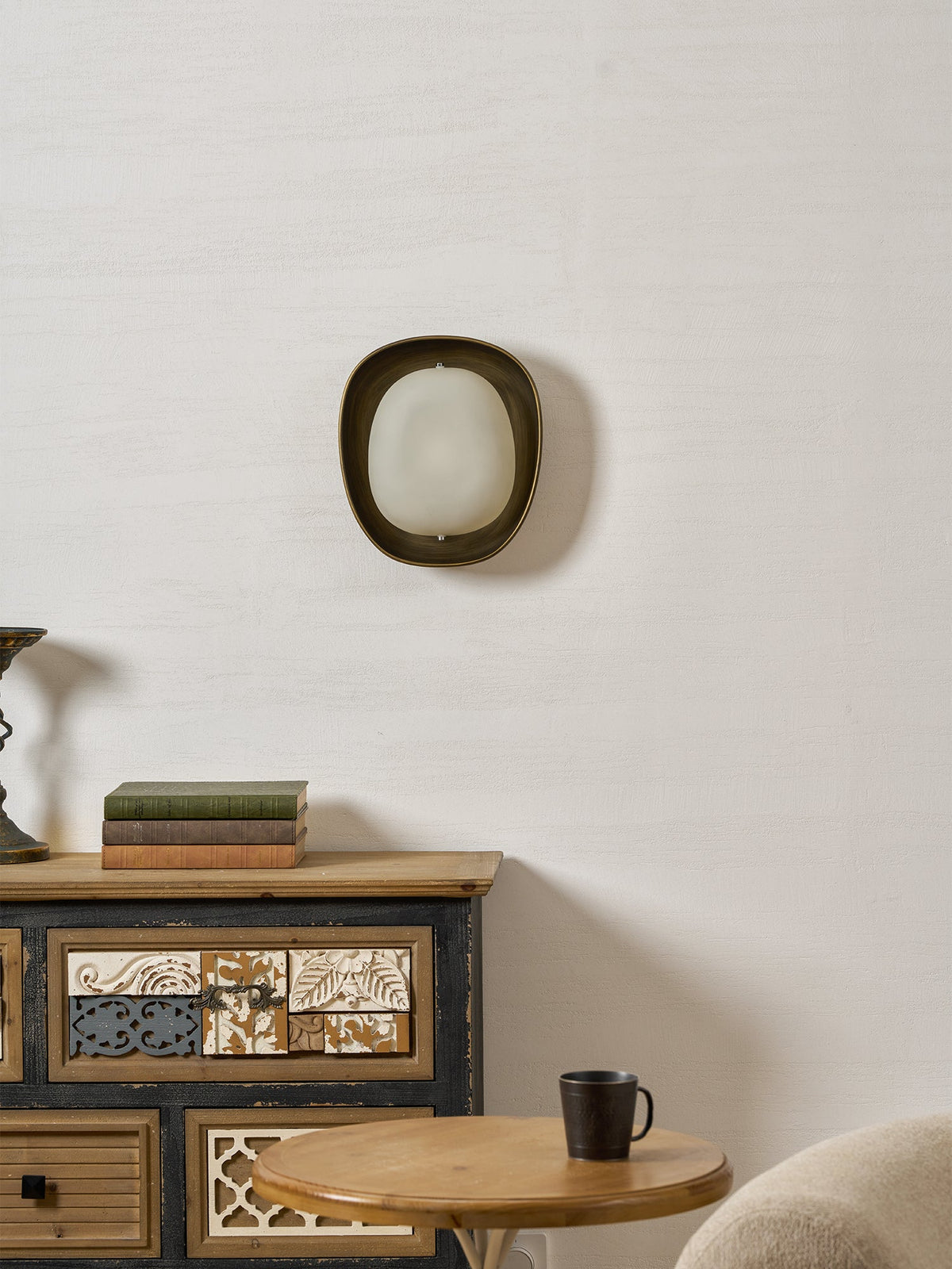 A small Clérachic wall light hangs on a pale wall above a painted sideboard, a round wooden table and a coffee mug.