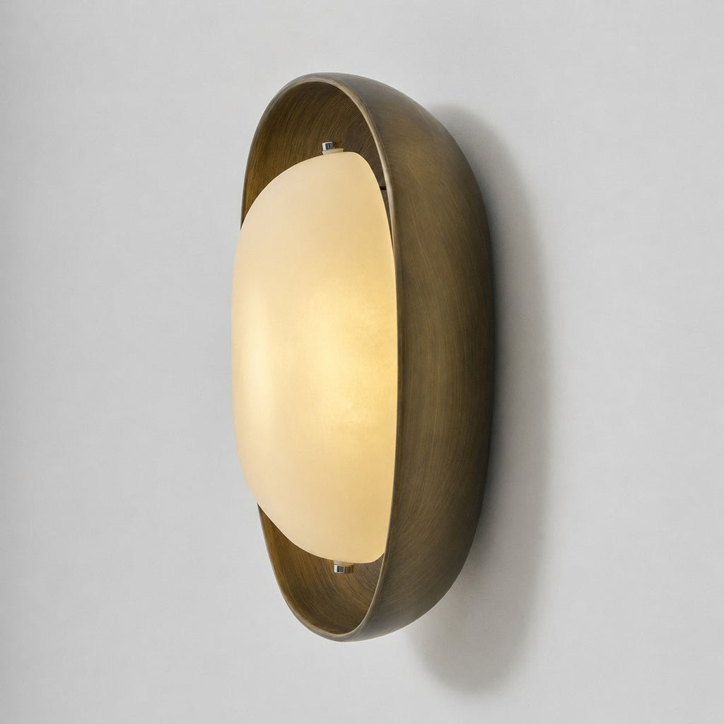 The frosted shade of this Clérachic wall light glows warm in a deep bronze bowl, seen from the side on a pale grey backdrop.