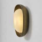 The frosted shade of this Clérachic wall light glows warm in a deep bronze bowl, seen from the side on a pale grey backdrop.