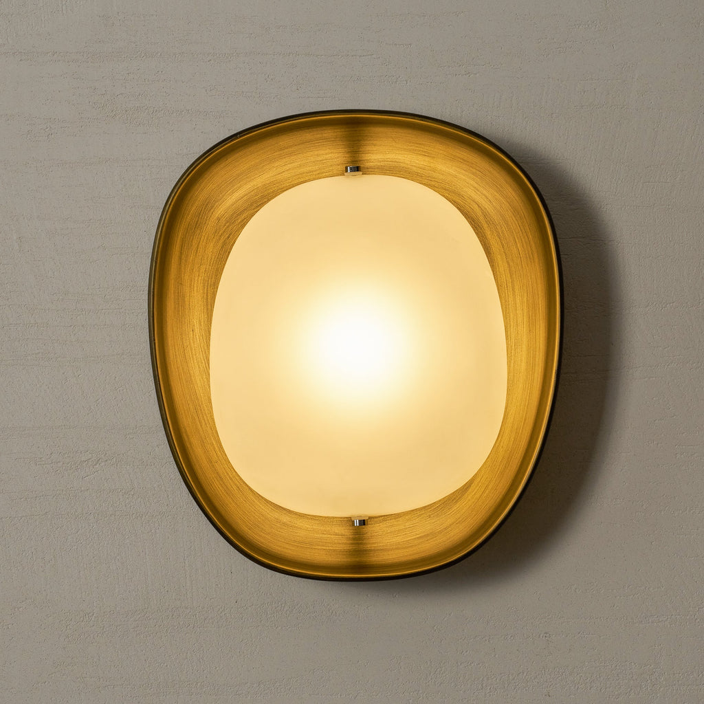 A lit Clérachic wall light glows gold on a grey plaster wall, with the bronze rim lit warm from inside.