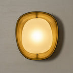A lit Clérachic wall light glows gold on a grey plaster wall, with the bronze rim lit warm from inside.