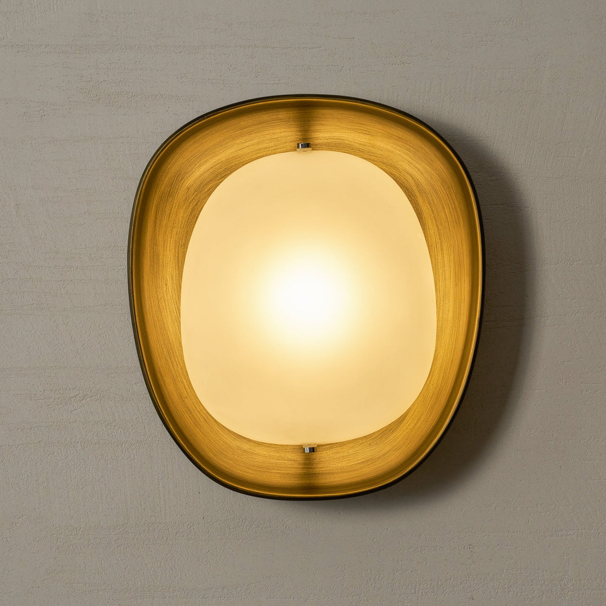 A lit Clérachic wall light glows gold on a grey plaster wall, with the bronze rim lit warm from inside.