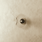Clérachic wall light with a free-form pale glass shade and a dark glass centre disc, switched off on a limewashed wall.