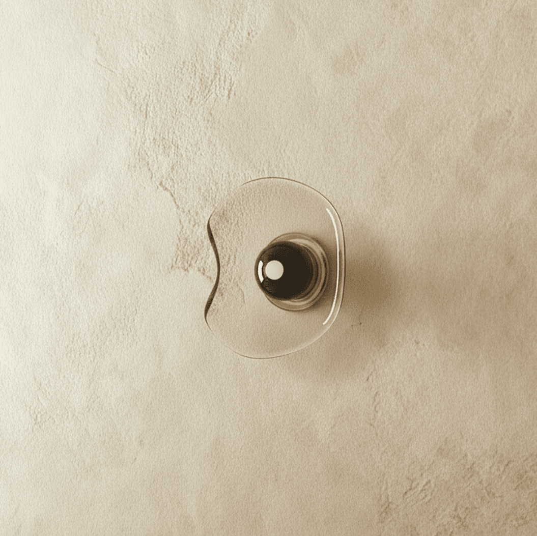 Clérachic wall light with a free-form pale glass shade and a dark glass centre disc, switched off on a limewashed wall.