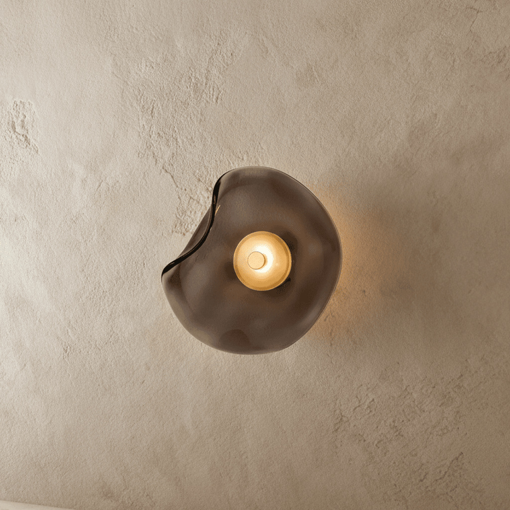 Clérachic wall light with a free-form smoky grey glass shade, its centre disc glowing warm on a textured plaster wall.