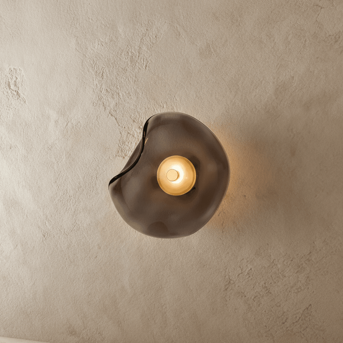 Clérachic wall light with a free-form smoky grey glass shade, its centre disc glowing warm on a textured plaster wall.