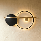 Clérachic black ring wall lamp with a black disc and a brass bar, its ring edge-lit with a warm glow on a limewash wall.