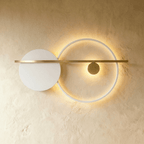 Clérachic white ring wall lamp with a white disc and a brass bar, its ring edge-lit with a warm glow on a limewash wall.