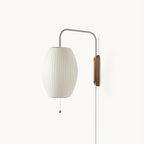 White ribbed oval shade on a curved steel arm with a walnut-toned wall bracket, shown on a plain white backdrop as a Clérachic plug-in wall light.