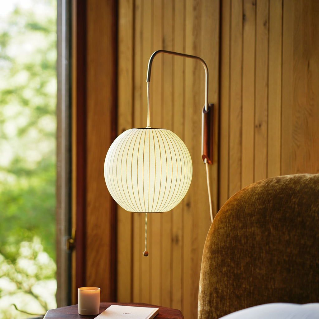 Round glowing shade on a Clérachic wall light against warm wood panelling, with a green window view and a lit candle below.