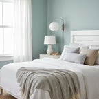 Round white Clérachic sconce on a pale blue wall above a white bedside table and lamp, beside a bed with a grey knitted throw.